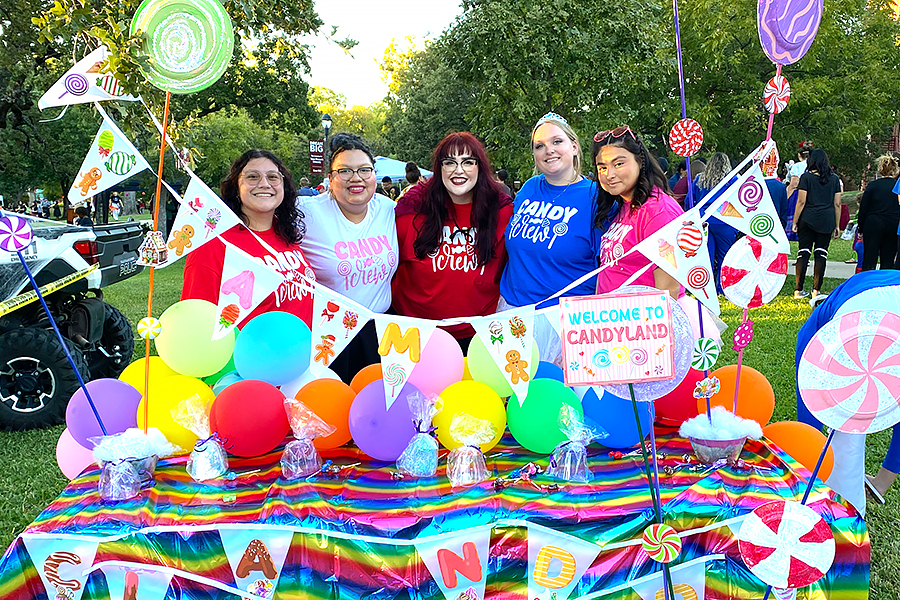 five TWU students in bright colored shirts stand behind a rainbow colored table at Boo at the U