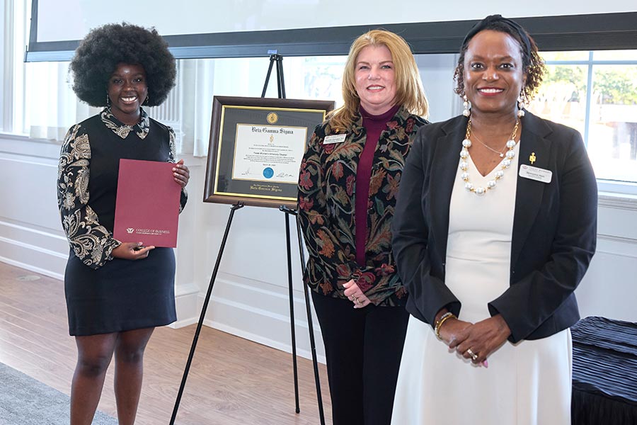 Theodora Sam-Brew holds a red folder, standing next to a Beta Gamma Sigma certificate and two faculty members
