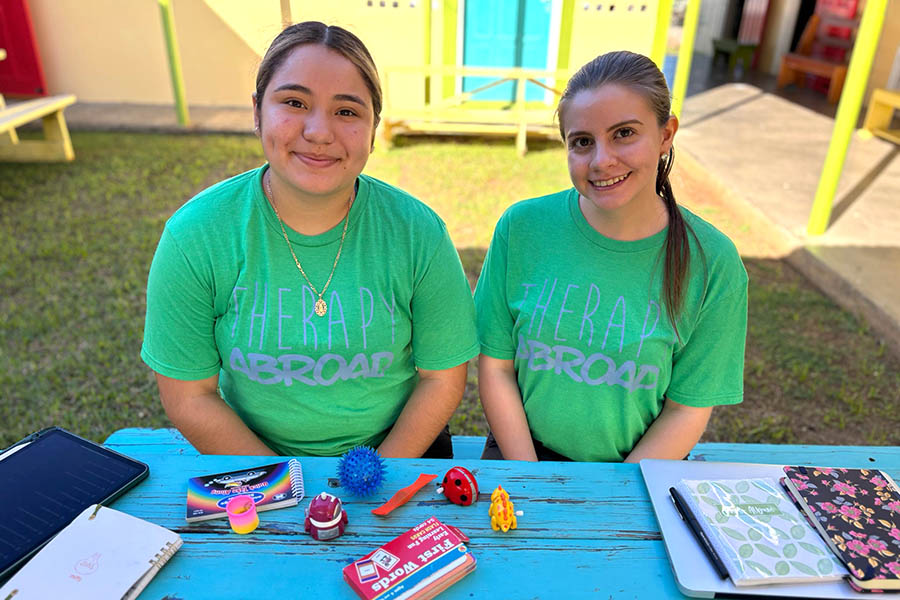 two students in green shirts sit at a table outside with SLp tools in front of them