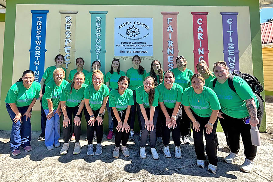 students and staff members in green shirts lined up in two rows stand in front of a sign