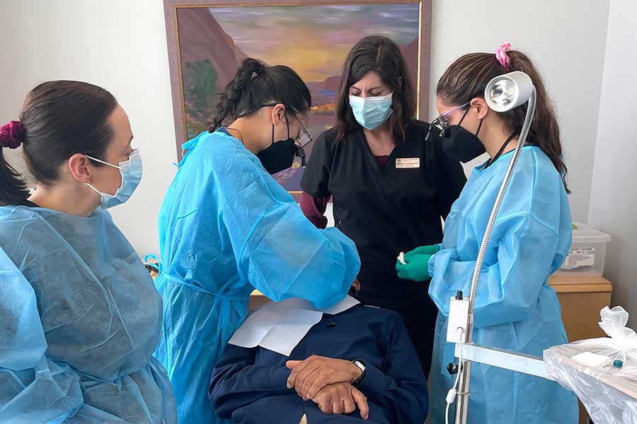 three DH students in blue scrubs care for patient on chair while staff member in black scrubs observes