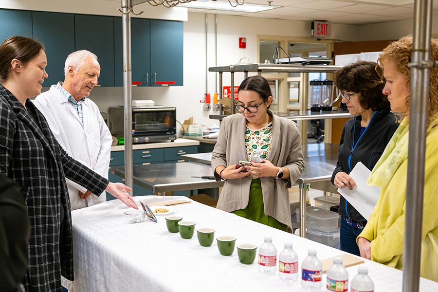 a food science student points to her food display on a kitchen counter to three judges