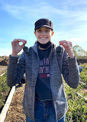 student wearing hat holding up 2 poblano peppers in garden