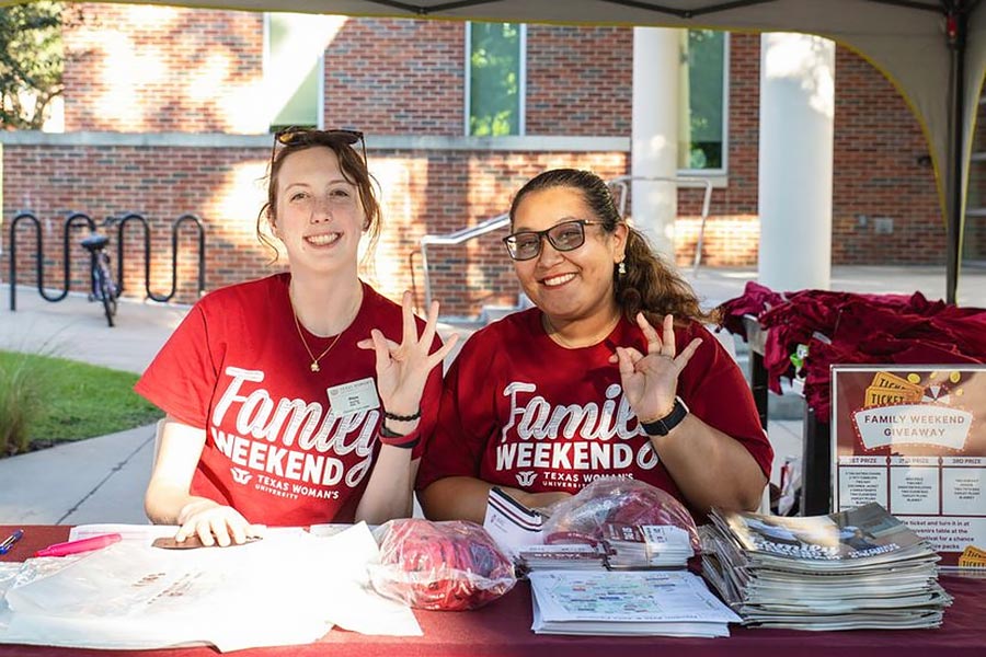 two students in red shirts sit behind a table holding up the university  hand sign
