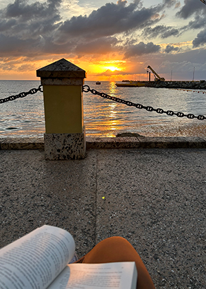 Kajilah Belardo in hat sits on beach chair on beach under an umbrellla