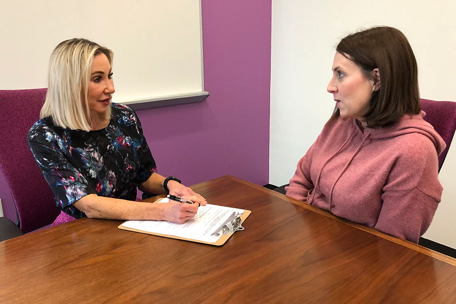 a IWH staff member writes on a clipboard seated on a table next to a female client during a consultation. 